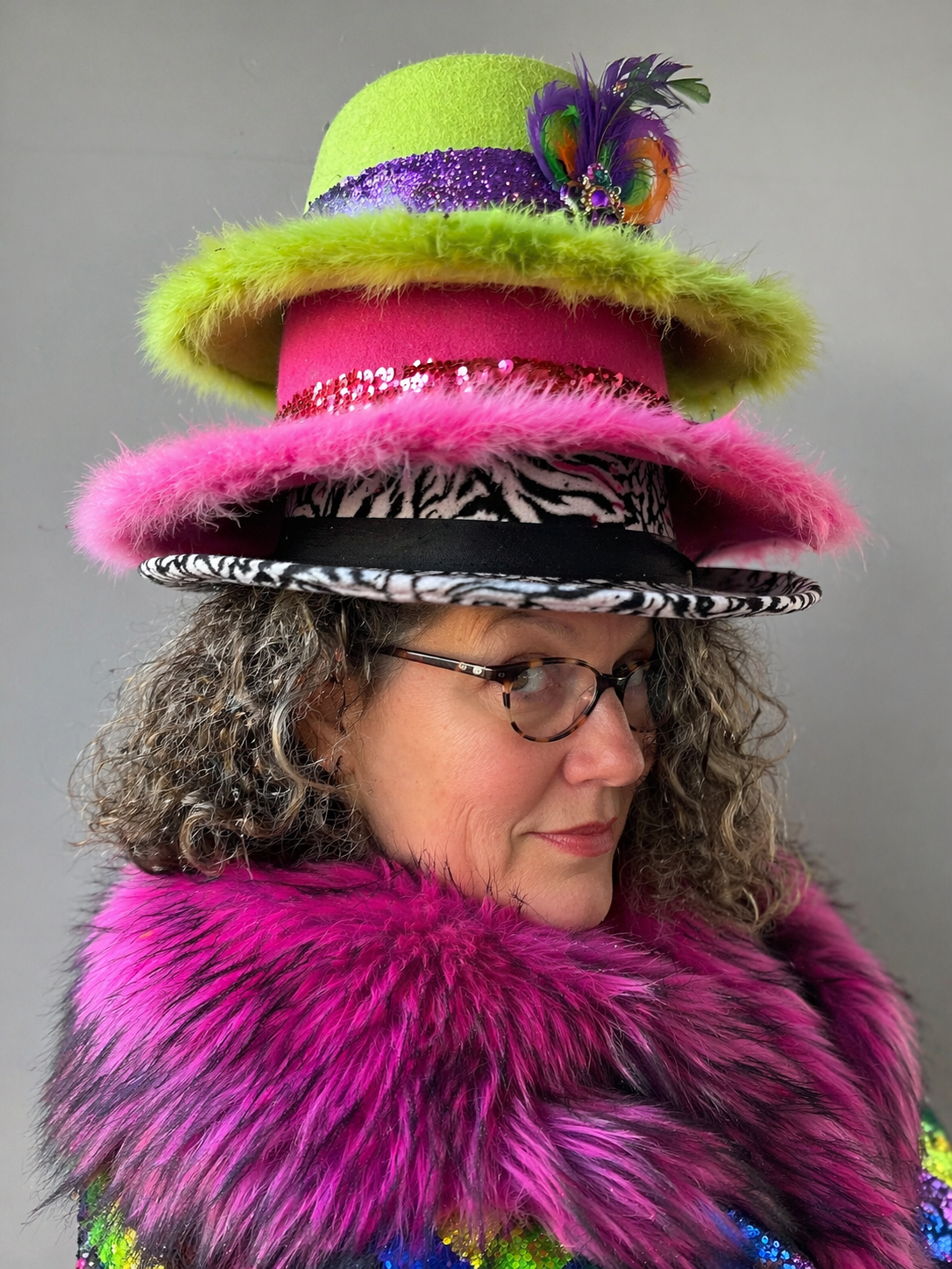 Vicki Peterson wearing three stacked hats — lime green, hot pink, and zebra-banded black — with a magenta fur collar